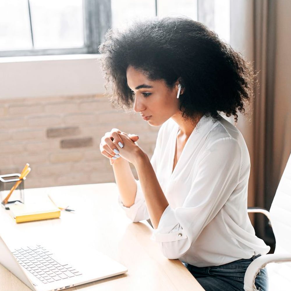 A woman with curly hair wearing a white blouse sits at a desk, looking intently at a laptop screen. She has wireless earbuds in and her hands are clasped under her chin. Office supplies are on the desk.