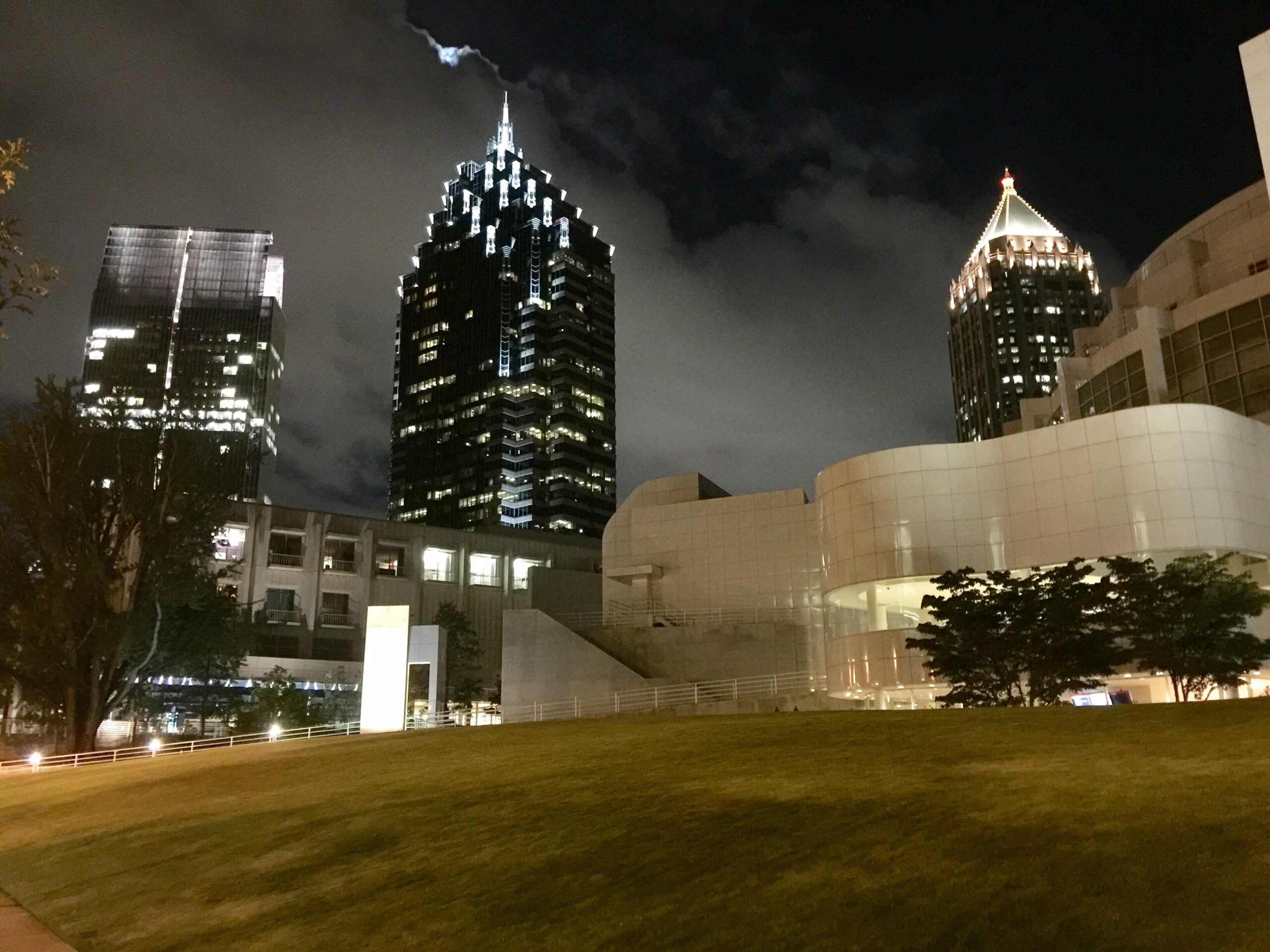A nighttime cityscape featuring modern skyscrapers with distinctive illuminated peaks behind a grassy lawn and a contemporary white building in the foreground. Clouds drift through the dark sky.
