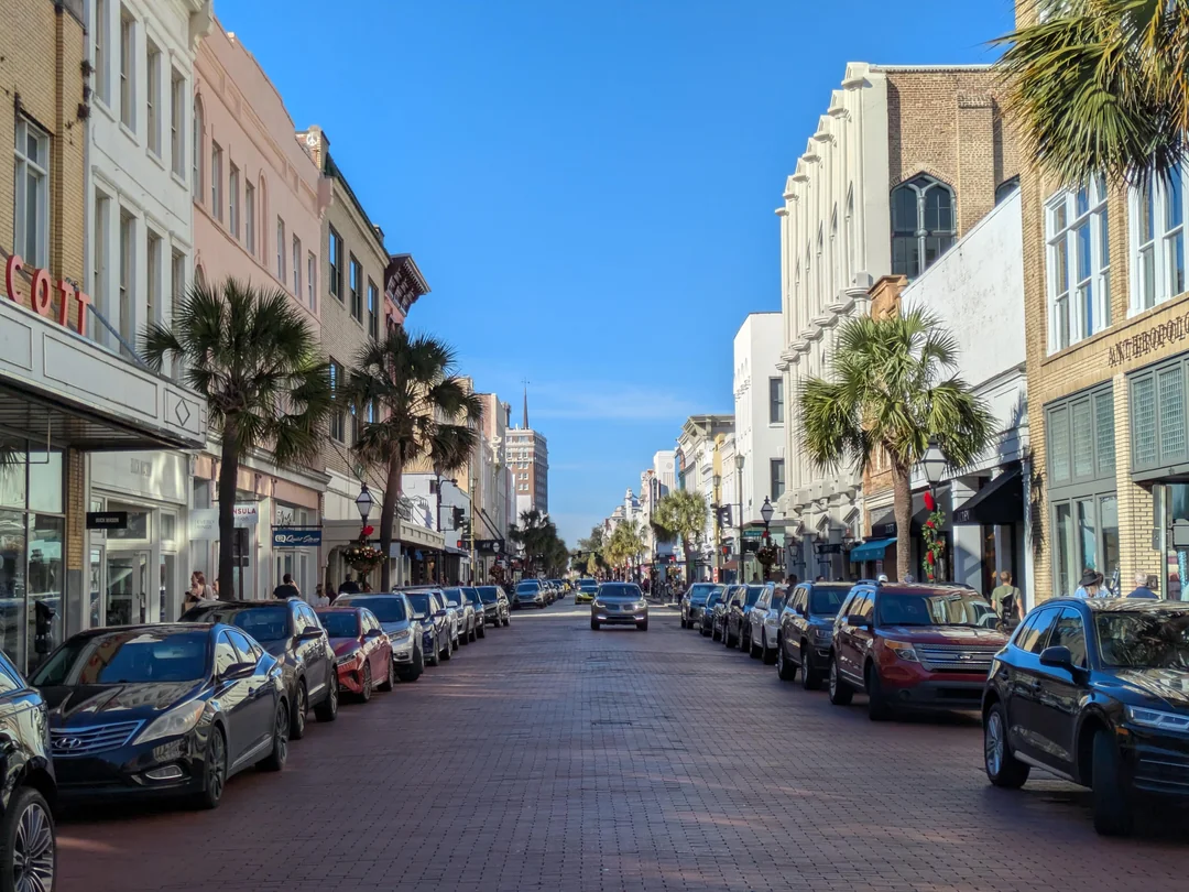 A brick-paved city street lined with parked cars and palm trees, bordered by historic, multi-story buildings on both sides under a clear blue sky.