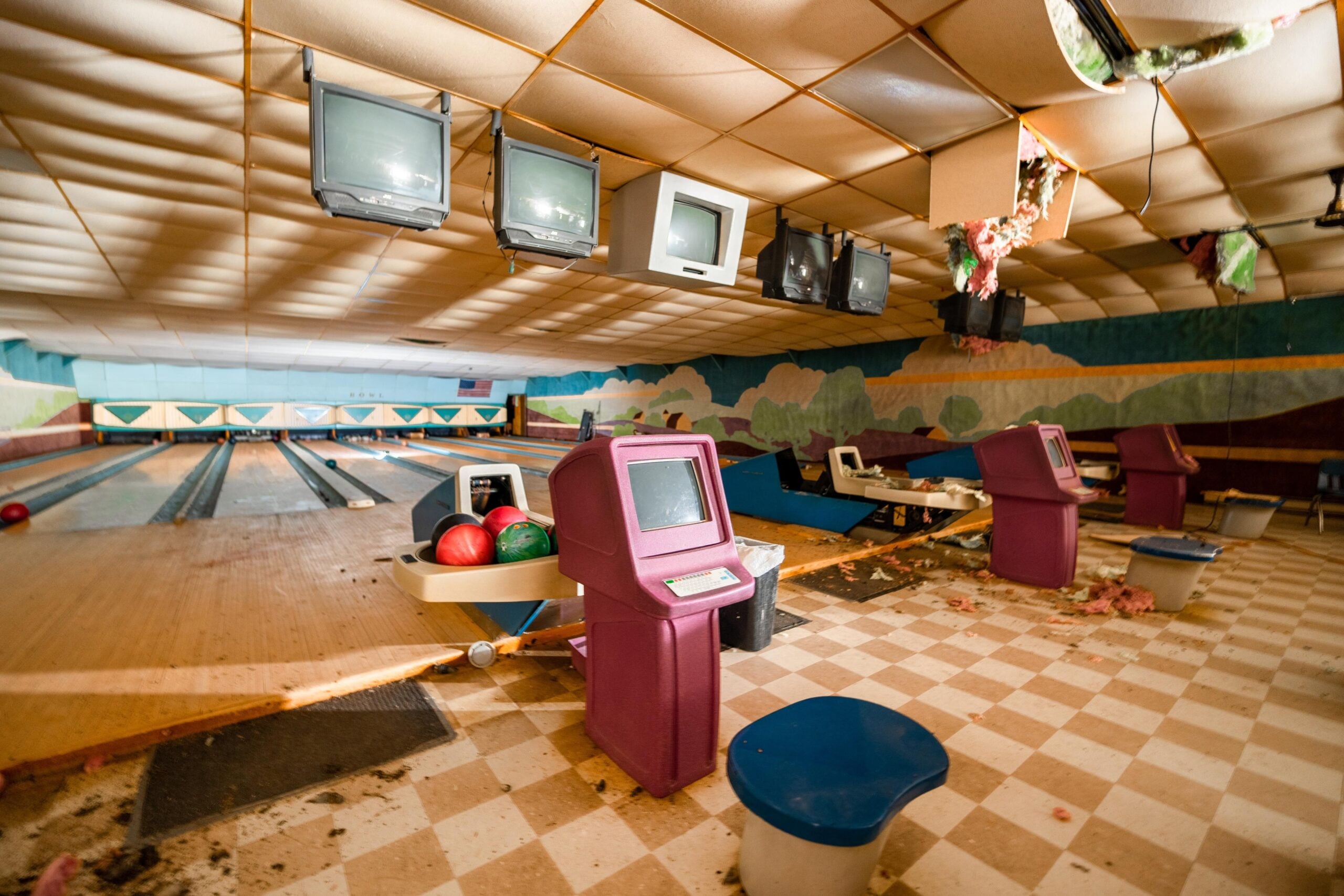 An abandoned bowling alley with dusty lanes, old CRT monitors, scattered bowling balls, and debris on a checkered floor. The ceiling has damage, and the area appears unused and neglected.