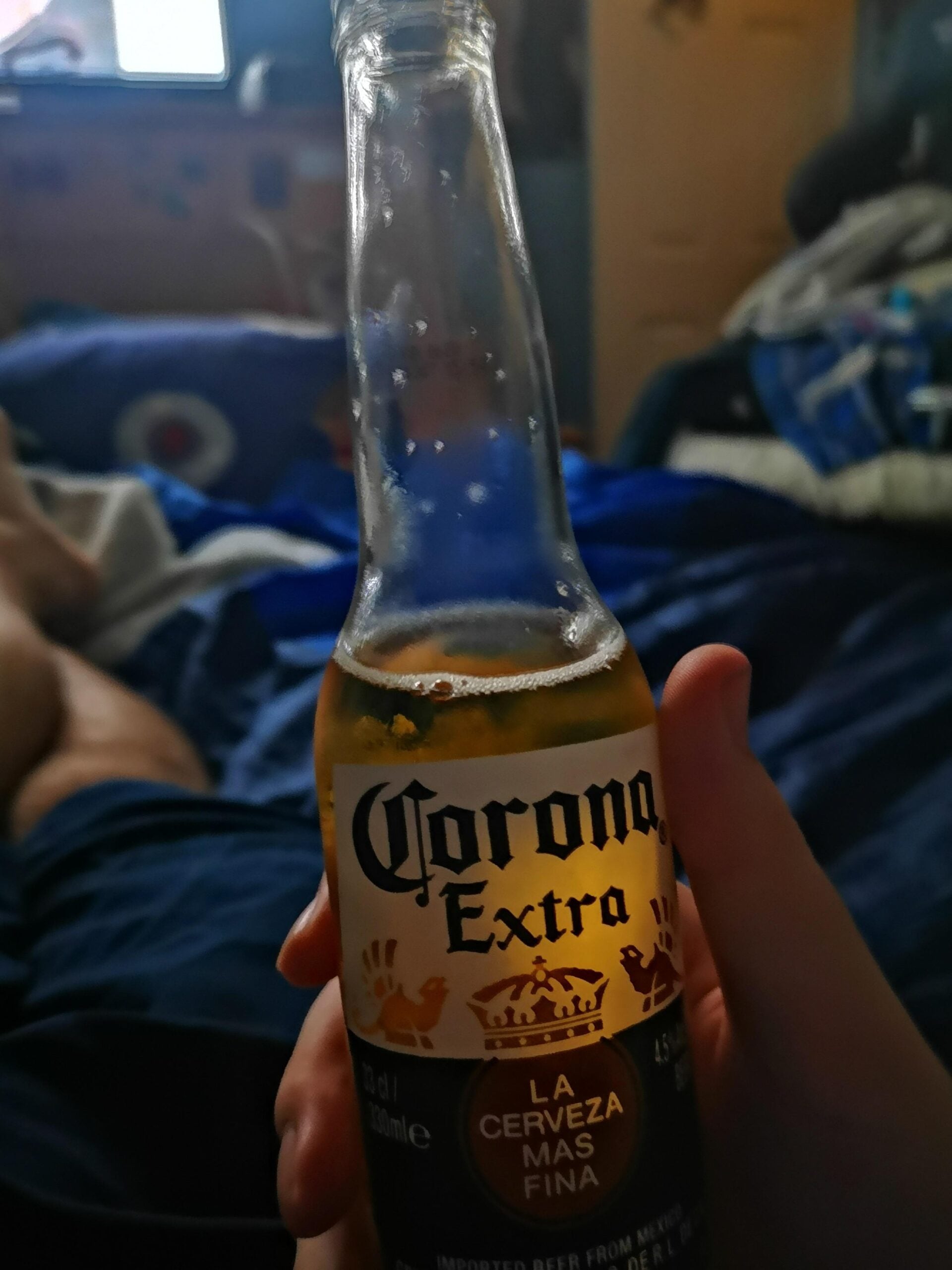 A person holds a Corona Extra beer bottle in their hand, sitting on a bed with blue sheets. The background shows scattered clothes and dimly lit room details.