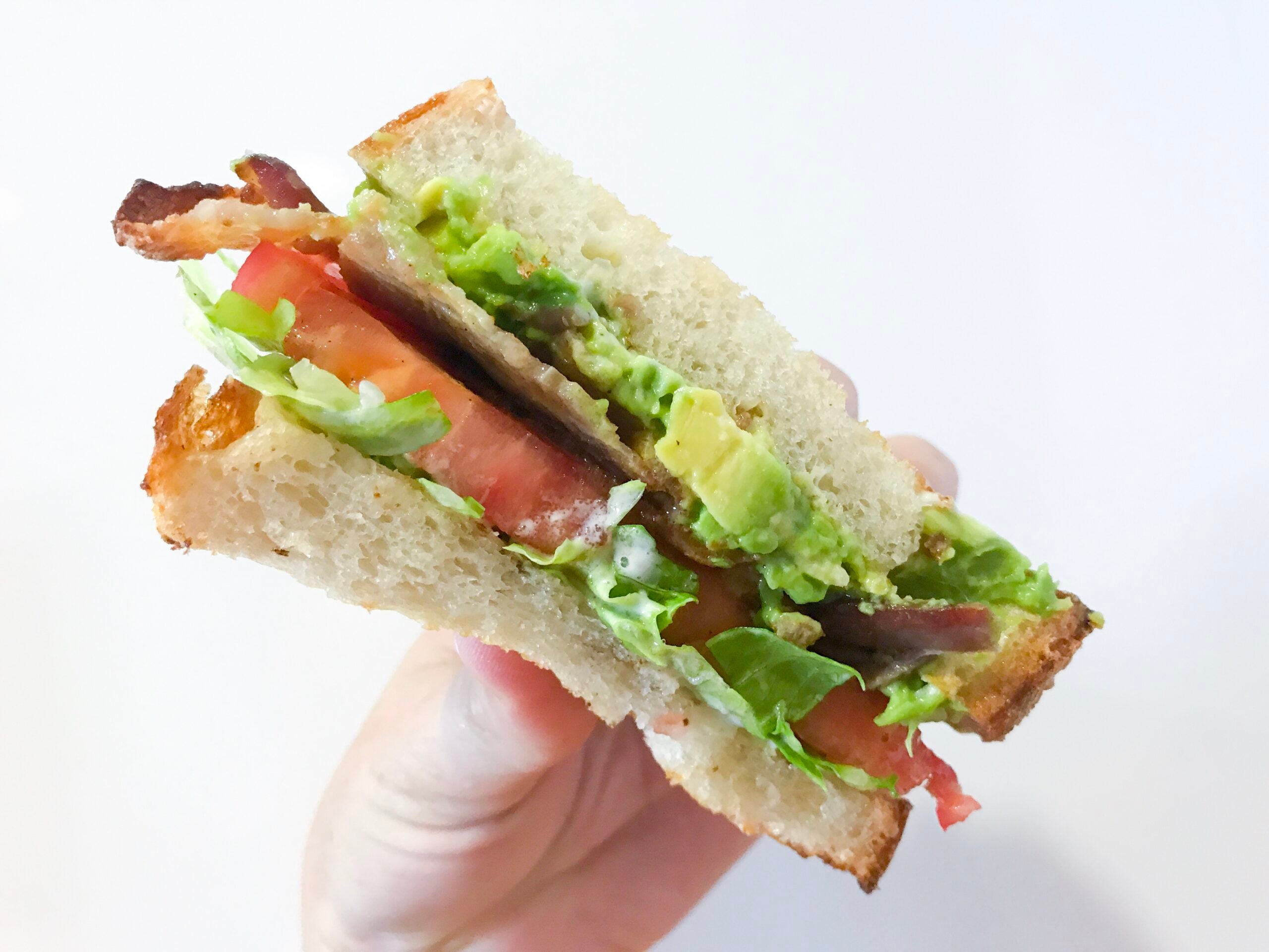 A hand holding a sandwich with layers of lettuce, tomato, bacon, and avocado between two slices of bread, against a plain white background.