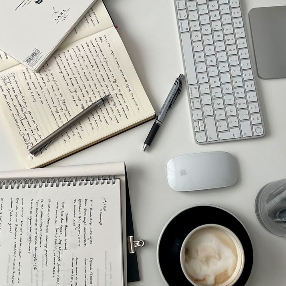 A workspace with an open notebook filled with handwritten notes, a pen, a spiral notebook, a wireless keyboard, a mouse, a closed laptop, and a cup of coffee on a saucer.