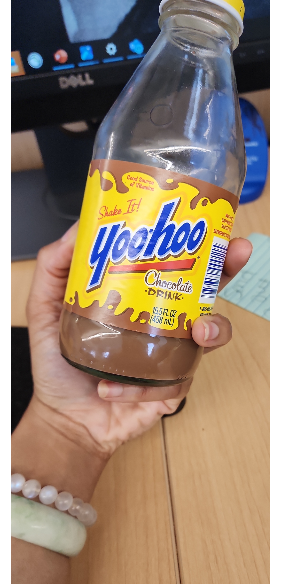 A hand holding a partially finished bottle of Yoo-hoo chocolate drink in front of a computer monitor on a wooden desk. The label is yellow with blue and brown text.