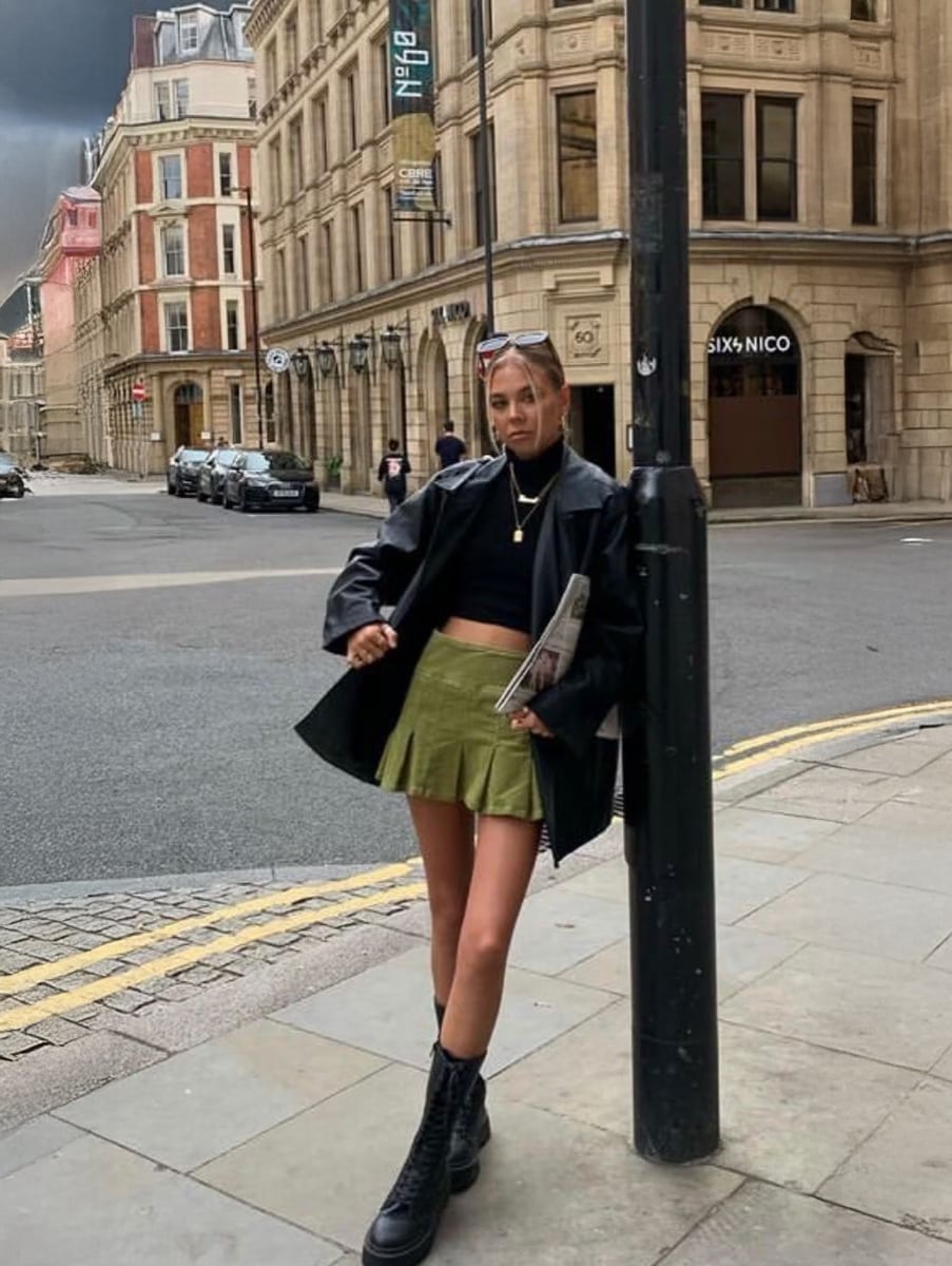 A woman wearing a black jacket, black crop top, olive-green skirt, and black boots stands on a city sidewalk holding a newspaper, leaning against a lamp post with historic buildings in the background.