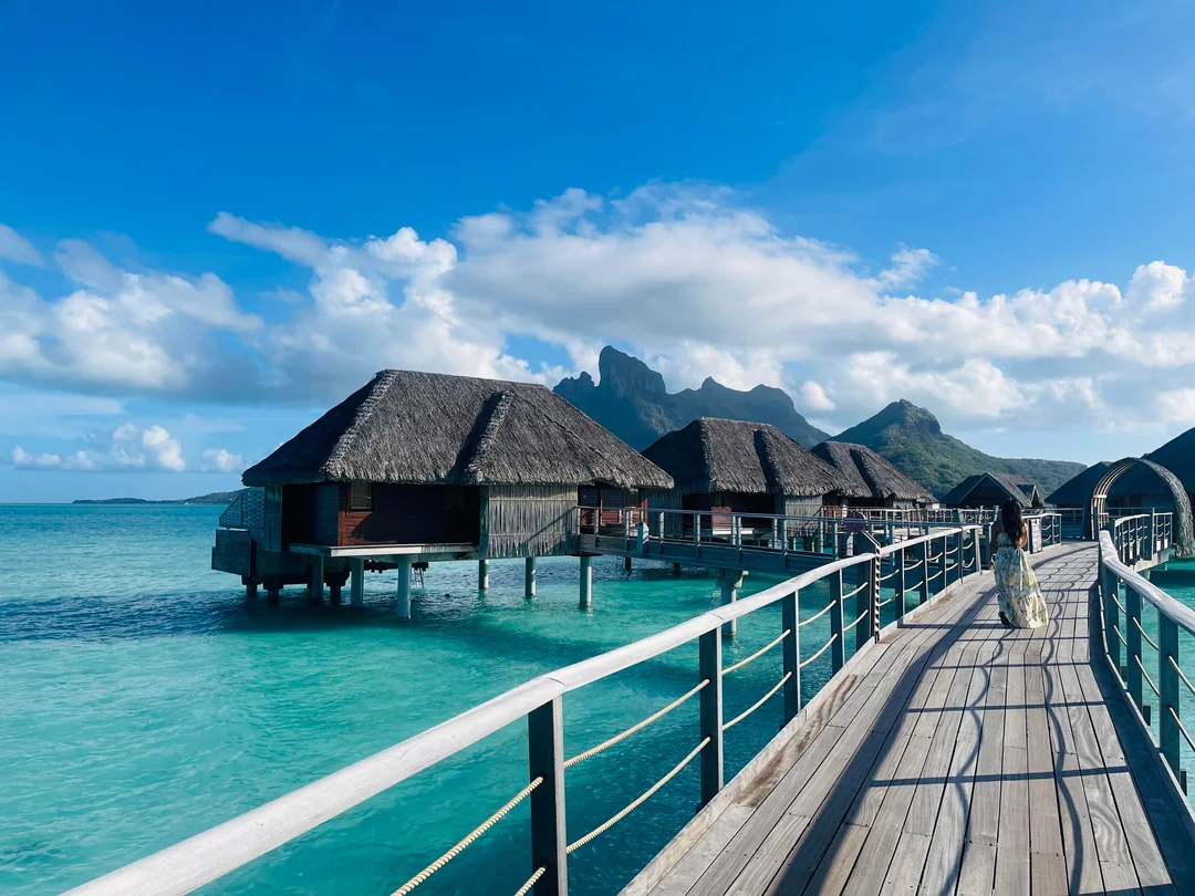 Overwater bungalows with thatched roofs sit above turquoise water, connected by a wooden walkway. Mountains and lush greenery rise in the background under a bright blue sky with scattered clouds.