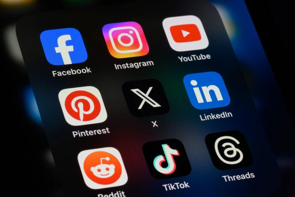 A smartphone screen displays various social media app icons, including Facebook, Instagram, YouTube, Pinterest, X, LinkedIn, Reddit, TikTok, and Threads, against a dark background.