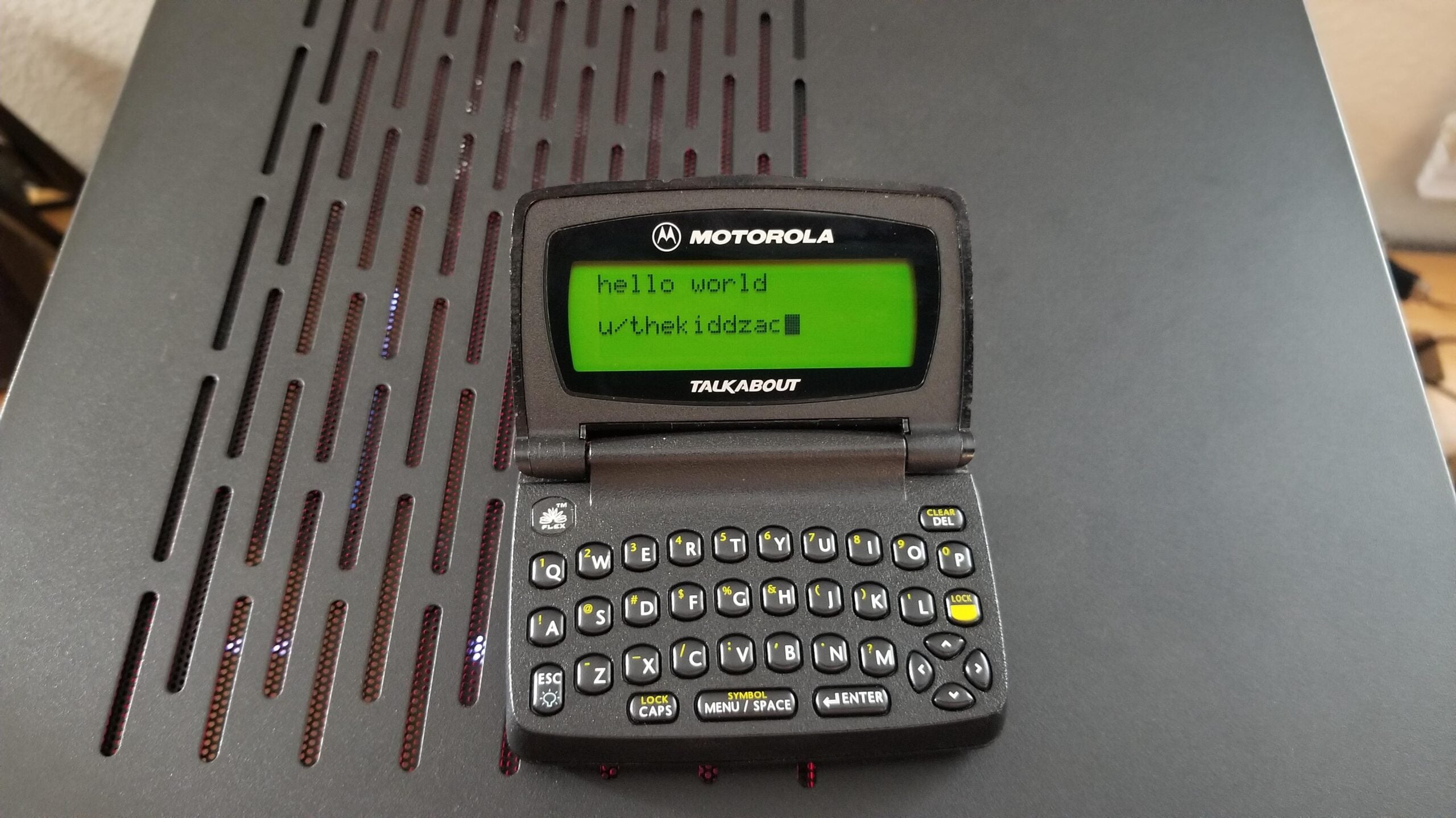 A Motorola Talkabout device with a small screen displays the text "hello world" and "u/thekiddzac." The device is placed on a black, vented surface.