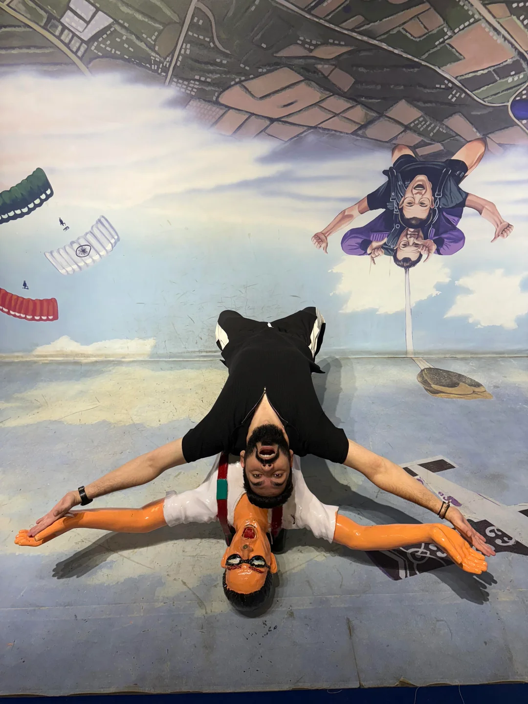 A man lies on his back, arms outstretched, mimicking the pose of a skydiving mannequin on the floor. The background is painted to look like the sky, with skydivers and parachutes, creating an optical illusion.