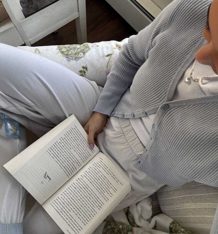 A person wearing light-colored, cozy clothes sits curled up on a chair, reading an open book in their lap. The scene looks comfortable and relaxed, with soft blankets and a peaceful atmosphere.