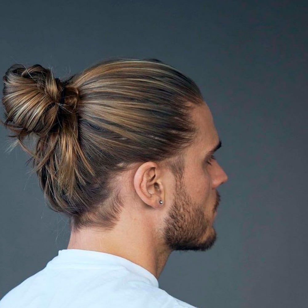 A man with long, light brown hair styled in a messy bun, wearing a white shirt and small earring, is shown in profile against a plain gray background.
