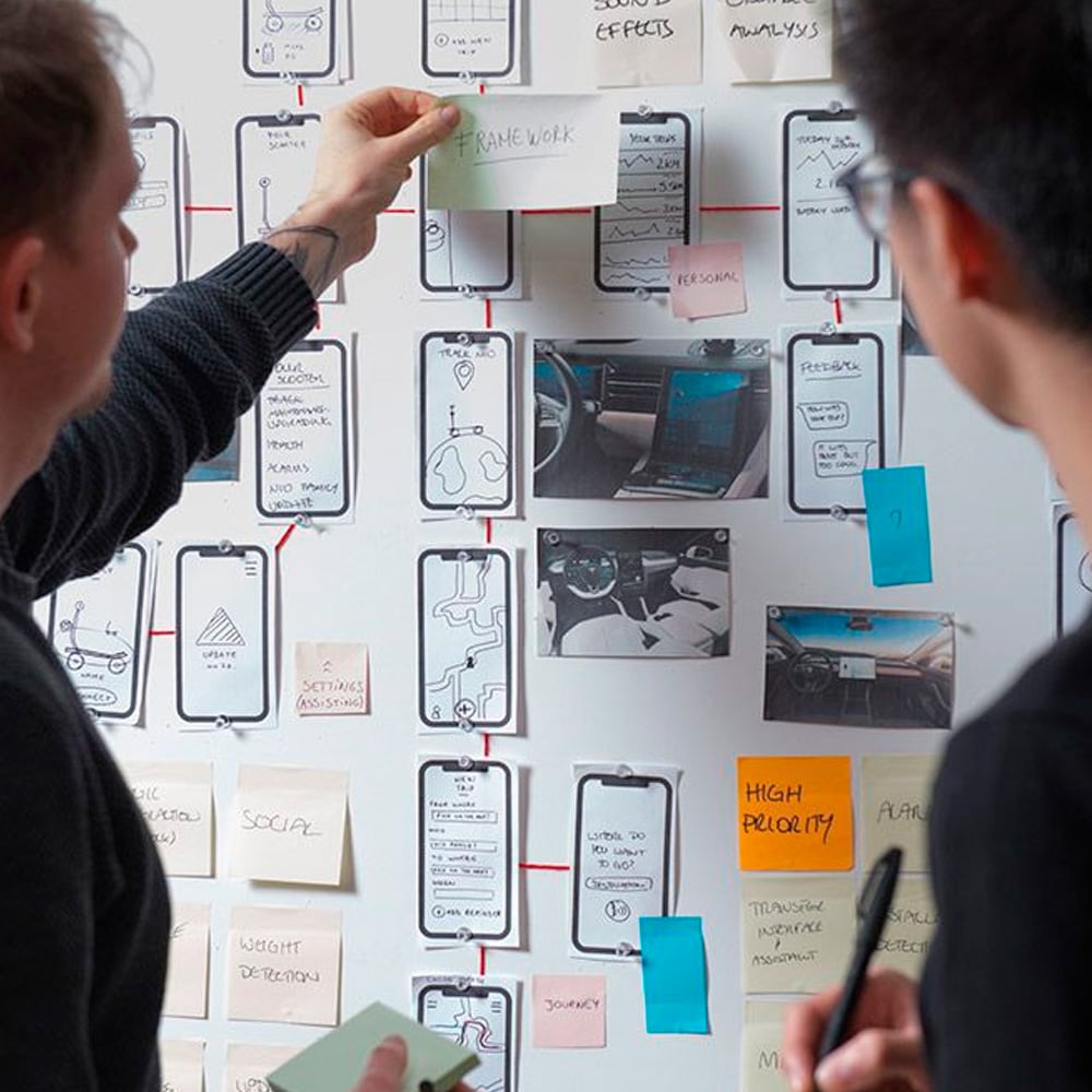 Two people discuss a wall covered with smartphone wireframes, photos, and colorful sticky notes, collaborating on a design project or app workflow using visual planning and notes.