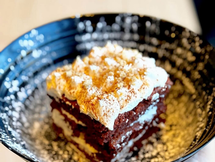 A slice of chocolate cake with layers of cream and topped with whipped cream and a dusting of cocoa powder, served on a black plate with powdered sugar sprinkled around.