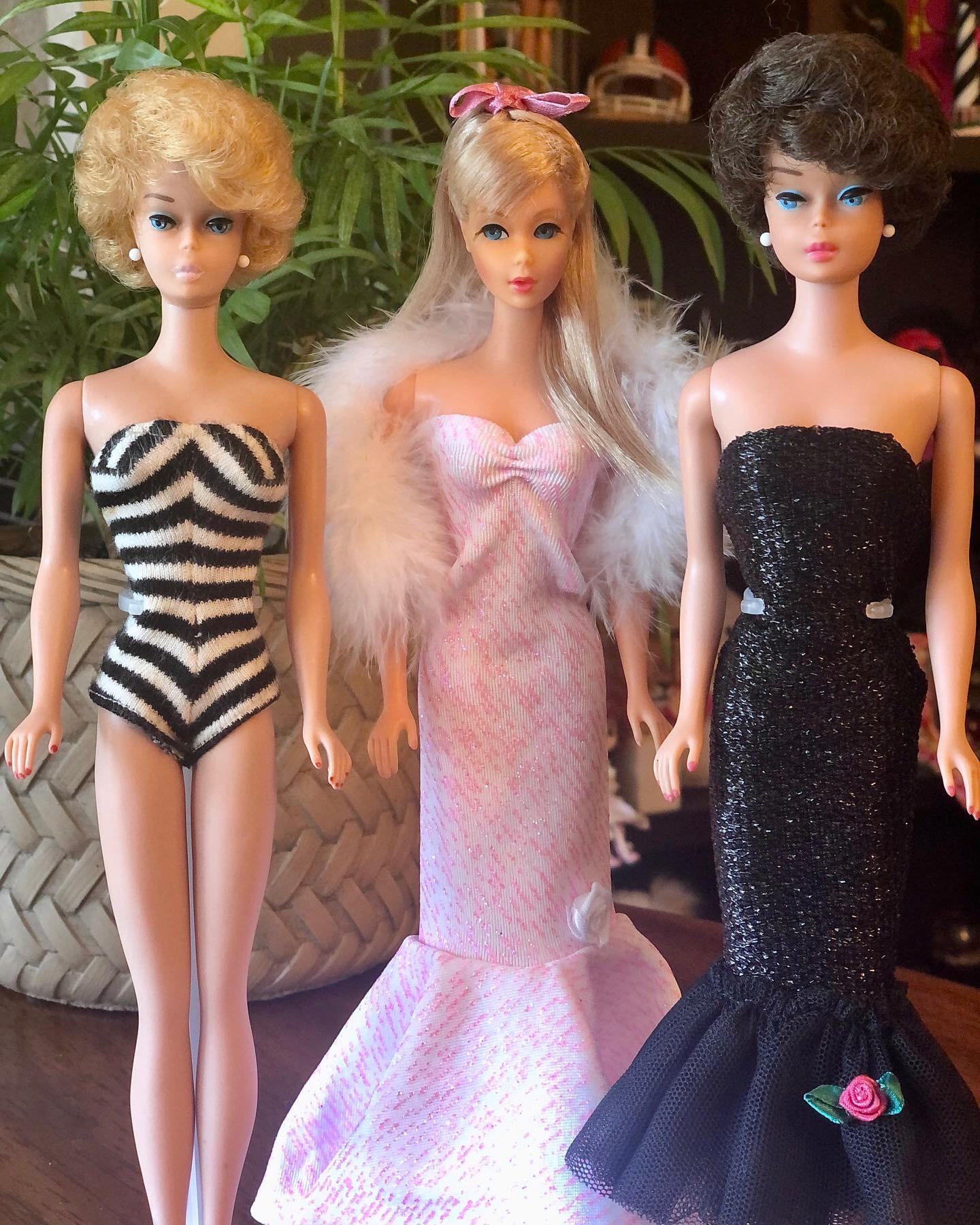 Three vintage Barbie dolls stand side by side. One wears a zebra-striped swimsuit, one a long pink gown with white feathers, and one a black strapless gown with a rose. A leafy plant is in the background.
