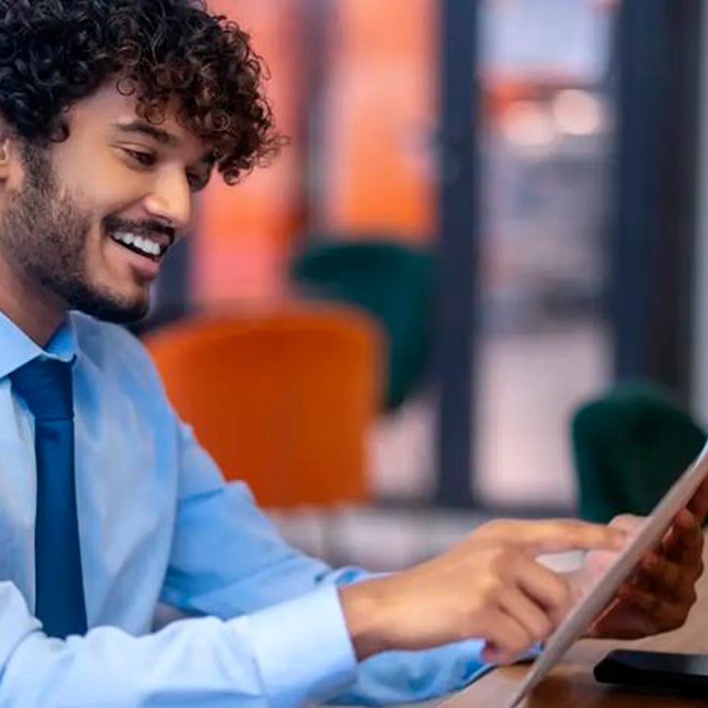 A smiling man in a blue shirt and tie is sitting at a desk and using a tablet, with colorful blurred office furniture in the background.
