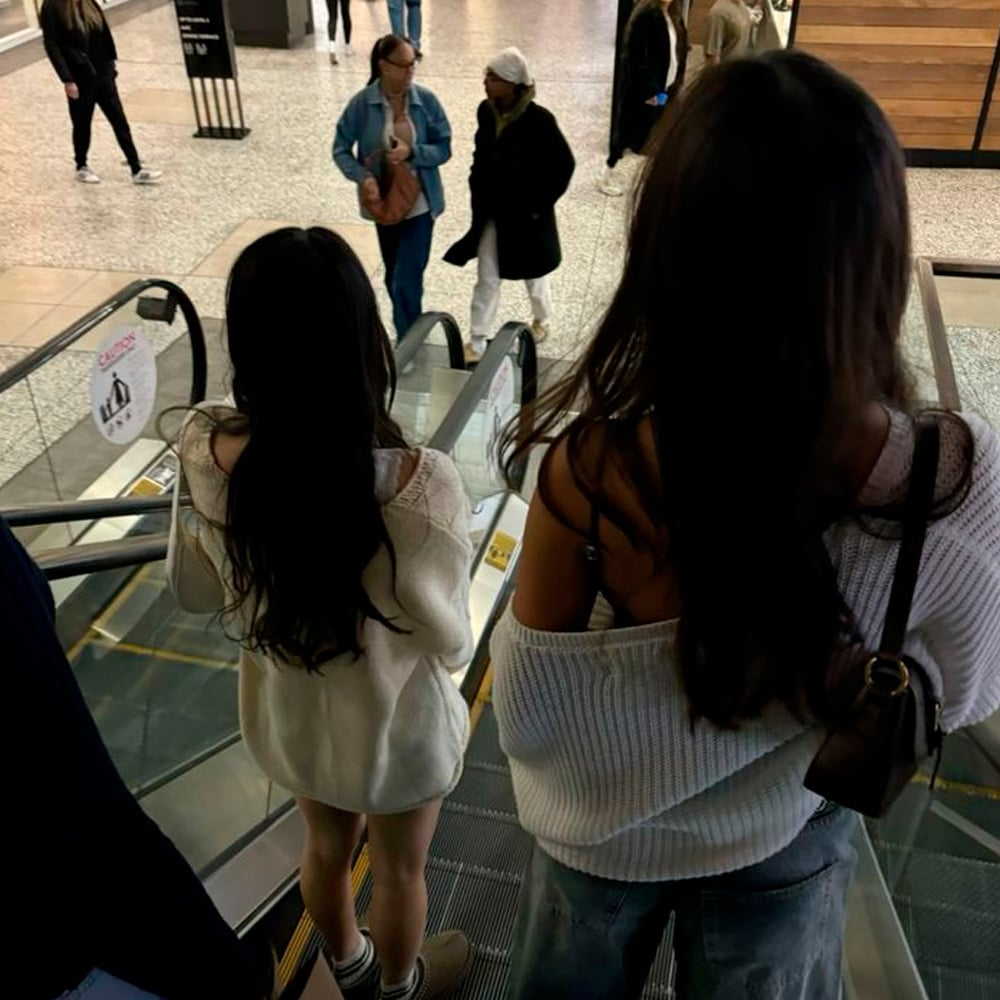 Two women with long dark hair stand on an escalator going down in a mall. One wears an oversized off-shoulder sweater, the other a white sweater and skirt. People walk in the background on a patterned floor.