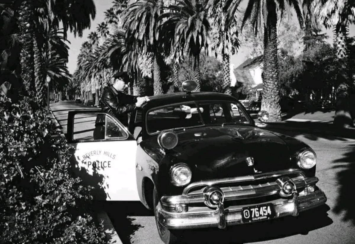 A vintage black-and-white photo shows a Beverly Hills police officer writing a ticket beside a classic police car parked on a palm tree-lined street.