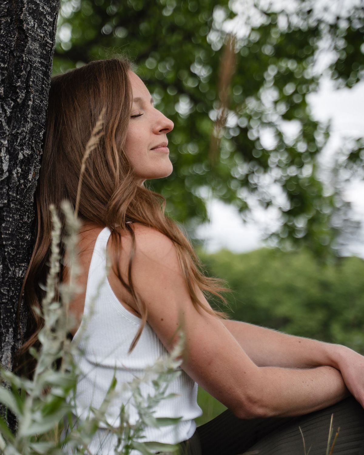 A woman with long brown hair, wearing a white tank top, sits outdoors with her eyes closed, leaning against a tree. Green foliage and grasses surround her, creating a peaceful, natural setting.