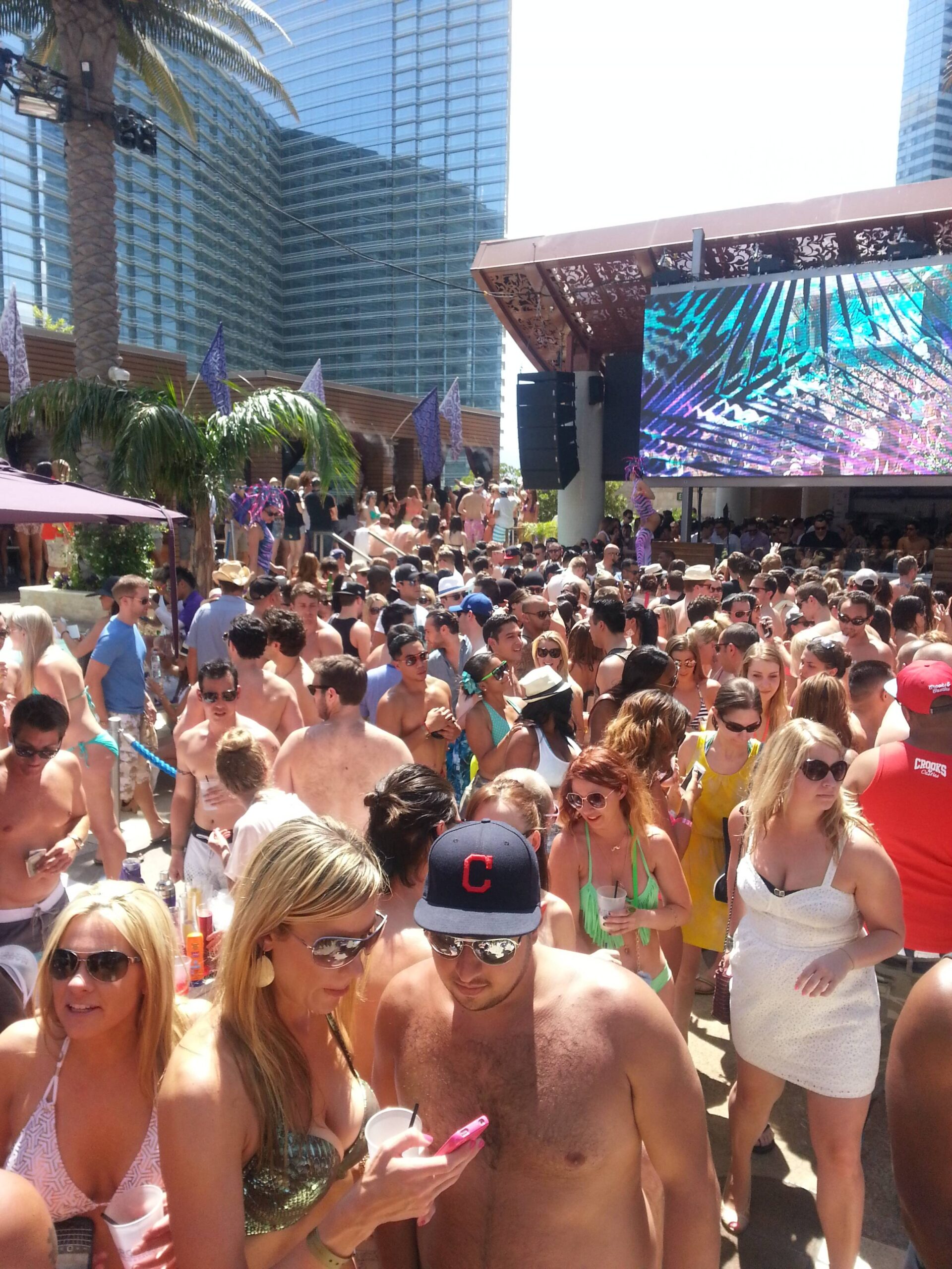 A large crowd of people in swimsuits gathers around a pool at a lively outdoor party with palm trees, drinks, and a large screen in the background. Bright sunlight and tall buildings surround the scene.