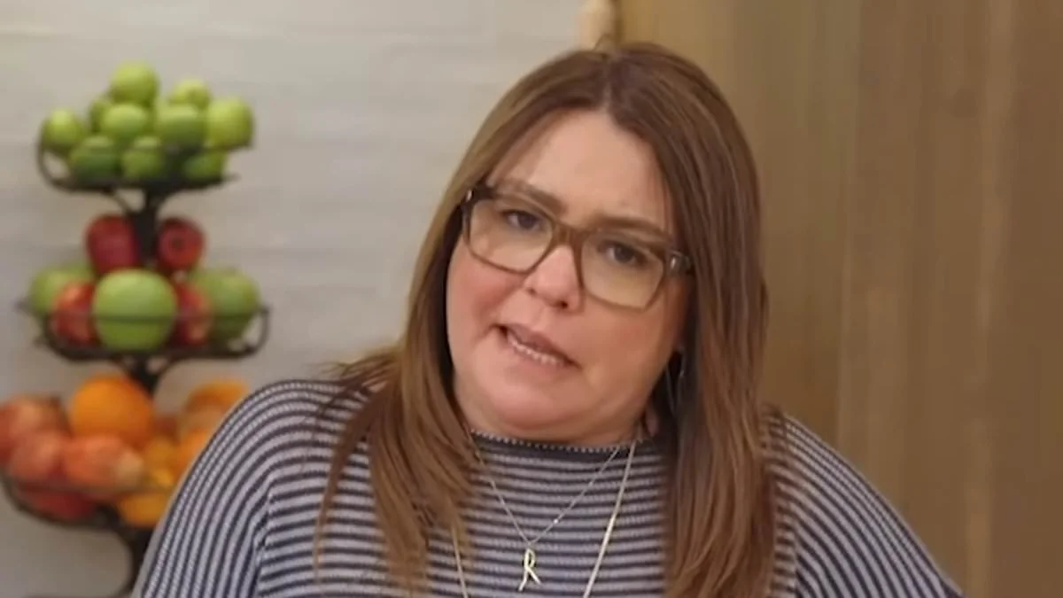 A woman with straight, light brown hair and glasses wears a striped shirt and an “R” necklace. She stands indoors in a kitchen with a fruit stand holding apples and oranges in the background.