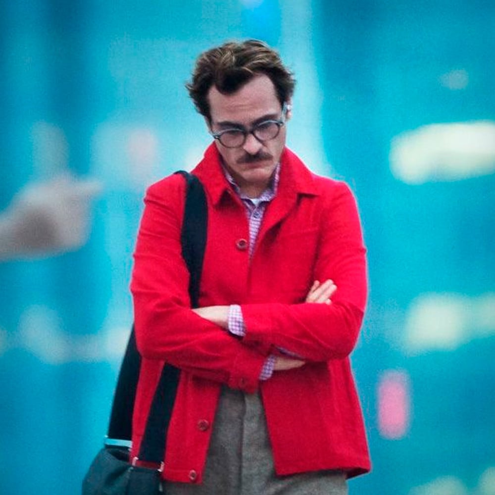 A man with glasses and a mustache, wearing a bright red jacket and carrying a shoulder bag, stands with his arms crossed and head down against a blurred blue city background.