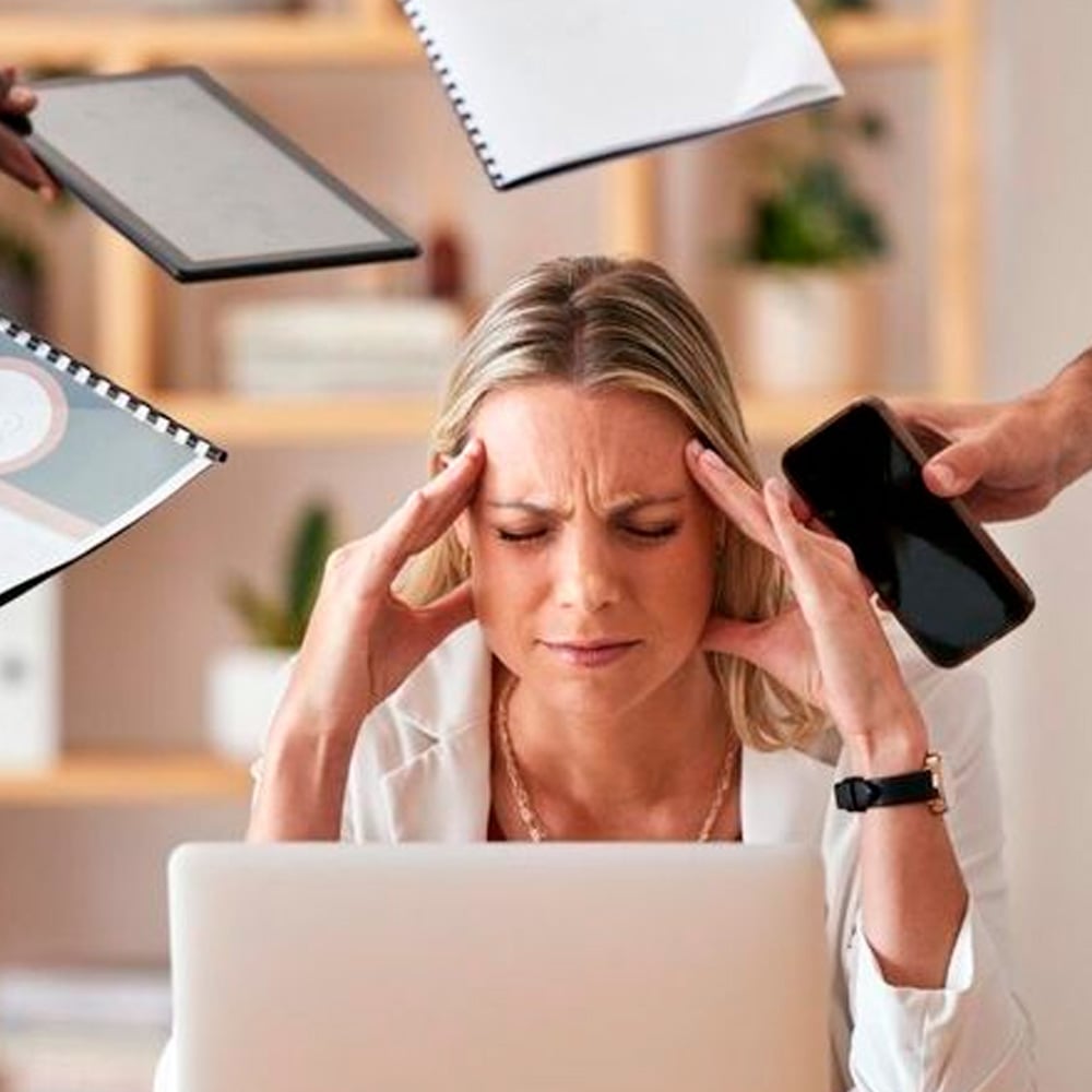 A stressed woman sits at a desk with her eyes closed, holding her temples, while people around her offer a phone, tablet, and documents, suggesting she is overwhelmed with work.