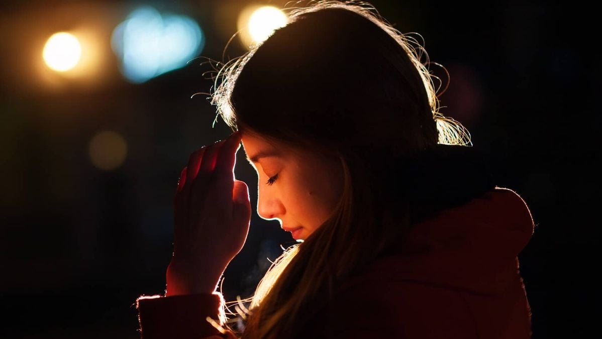A woman with long hair stands in profile at night, eyes closed, touching her forehead with one hand. She is illuminated by warm, dramatic lighting, with blurred lights glowing softly in the background.