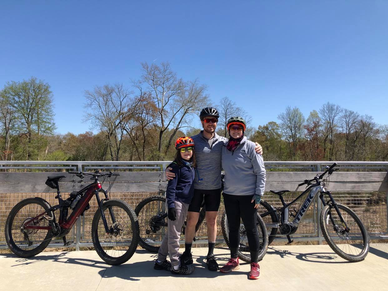 Three people wearing helmets and casual biking clothes stand together on a bridge, smiling, with two bicycles parked nearby. Trees with spring foliage and a clear blue sky are in the background.