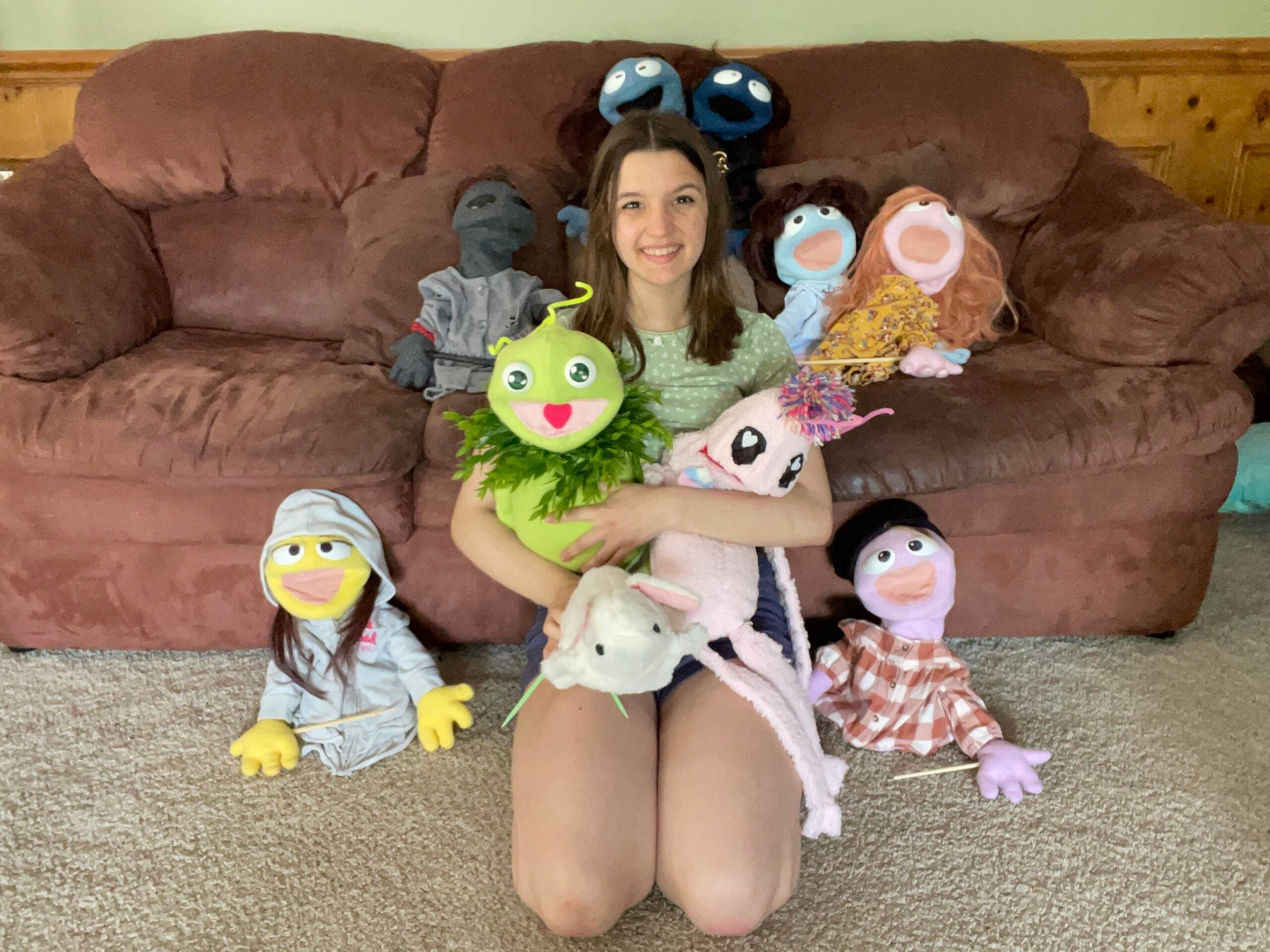 A young woman sits on the floor in front of a brown couch, smiling and holding several colorful puppets and stuffed animals, with more puppets arranged around her on the floor and couch.