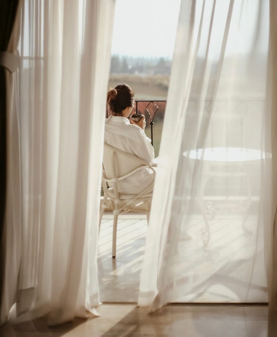 A person in a white robe sits on a balcony chair, holding a cup and looking out at the view. Sheer white curtains frame the scene, letting in soft natural light. A small table is next to the person.