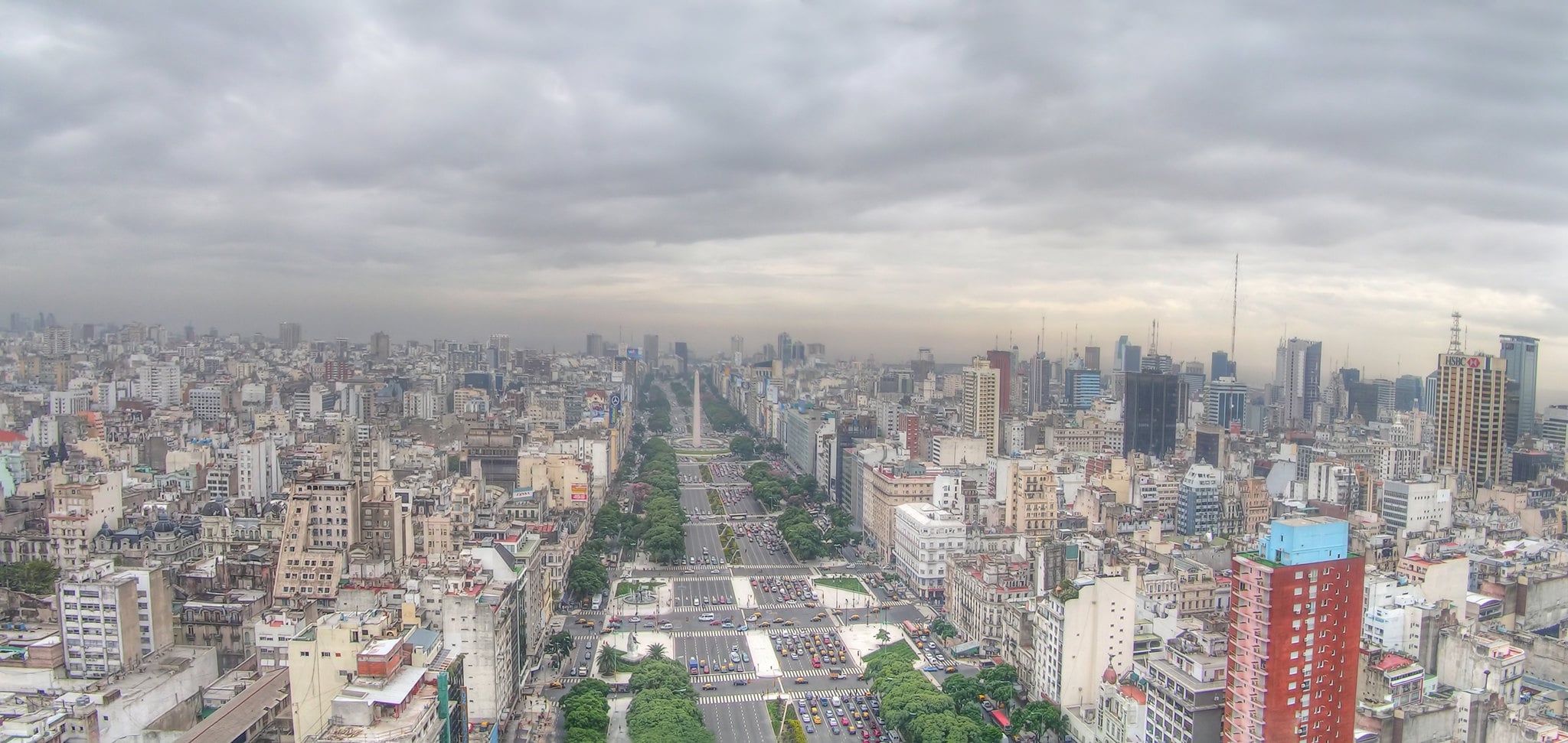 Aerial view of a wide city avenue lined with trees and surrounded by tall buildings under a cloudy sky in an urban area. Traffic is visible on the avenue below.