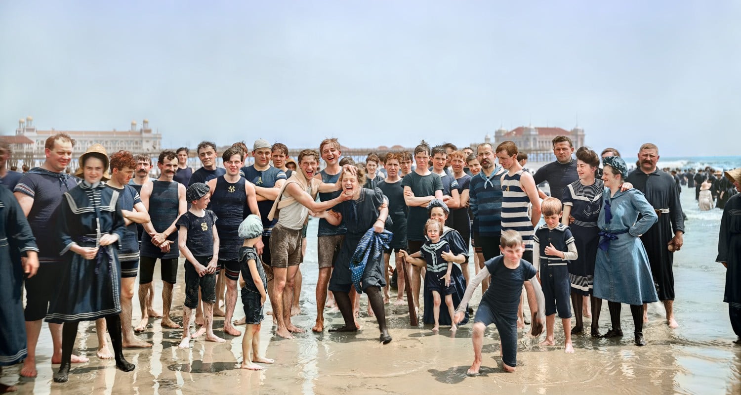 A large group of men, women, and children in vintage swimsuits stand together smiling and posing on a sandy beach, with waves and distant buildings in the background.