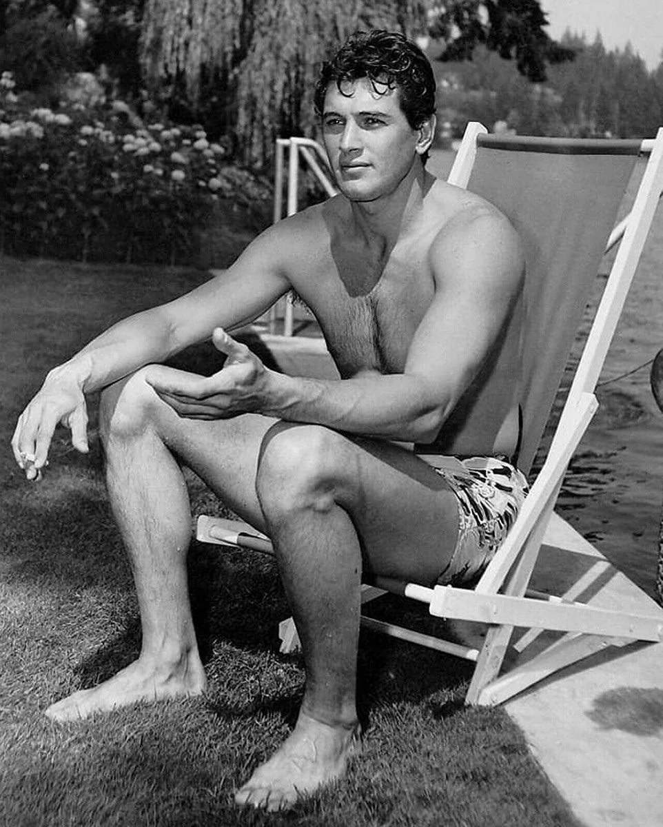 A man in patterned swim trunks sits on a folding lounge chair by a lake, gesturing with one hand. He is barefoot and shirtless, with short dark hair, and the background features grass, trees, and water.