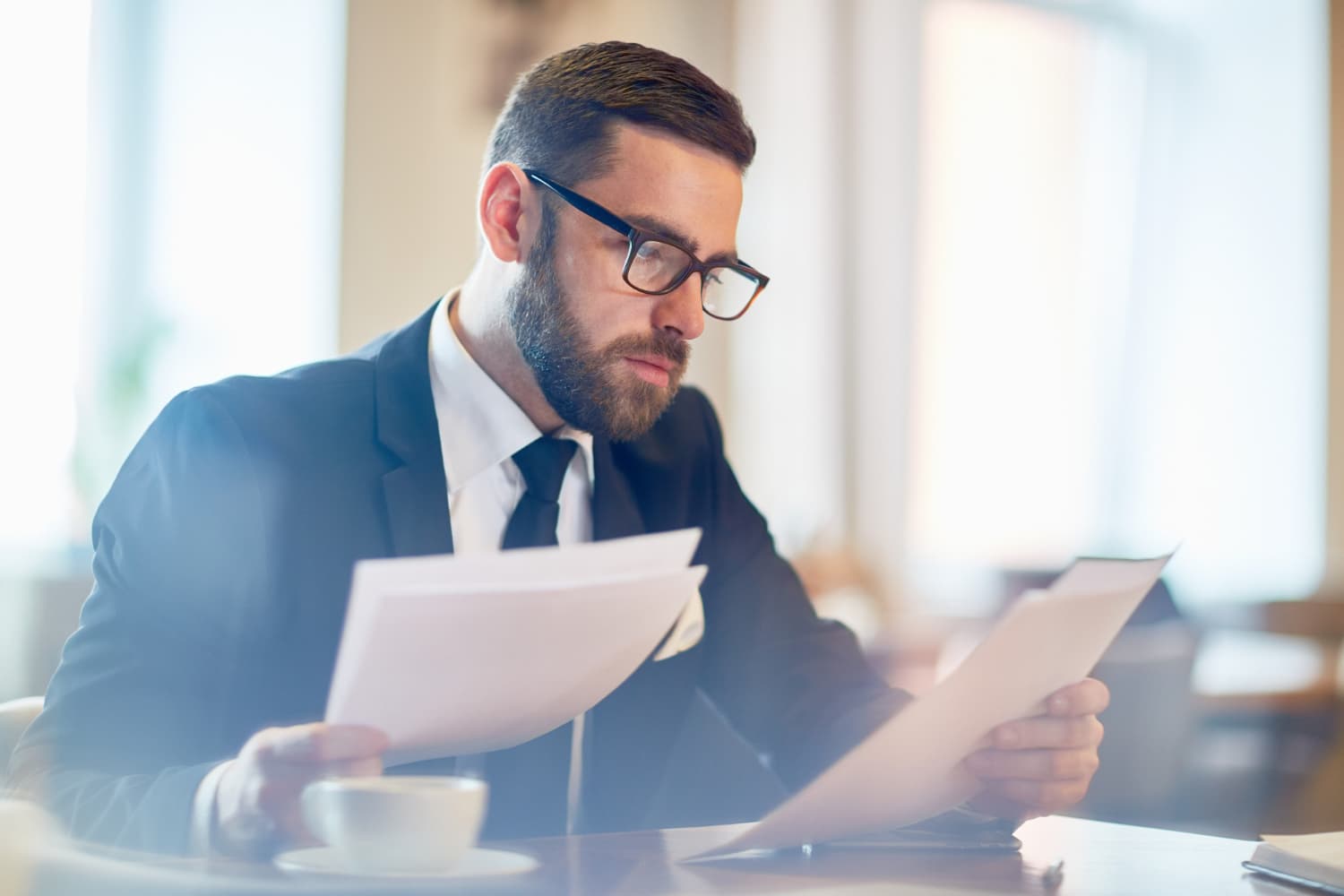 A man in a suit and glasses sits at a table, reading documents intently. He has a beard and is holding papers in both hands. A cup and saucer are on the table in front of him. The background is softly blurred.