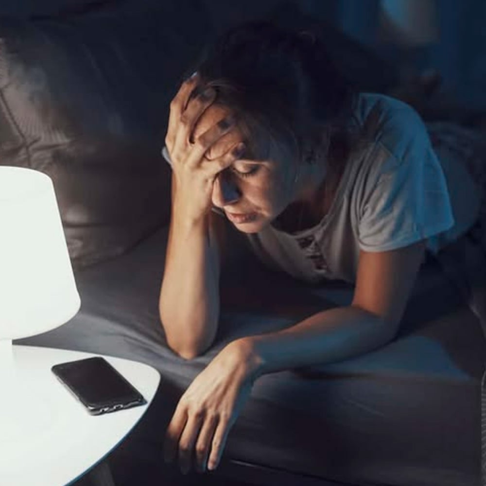 A woman sits on her bed at night, head resting in one hand, looking stressed or exhausted. A smartphone lies on a nearby nightstand, illuminated by a bedside lamp.