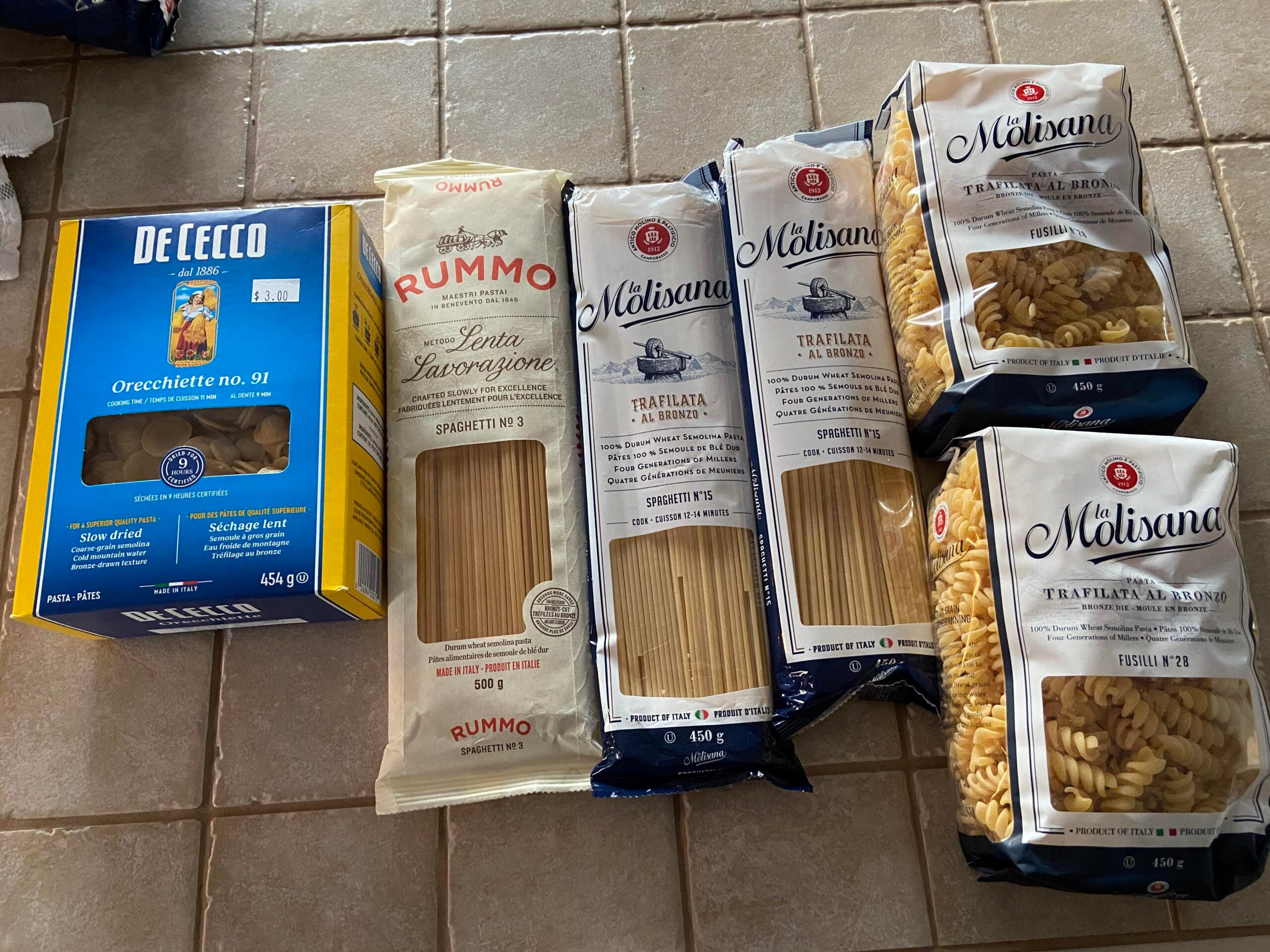 Five packages of Italian pasta, including De Cecco orecchiette, Rummo spaghetti, and three varieties of La Molisana pasta, are arranged on a beige tiled surface.