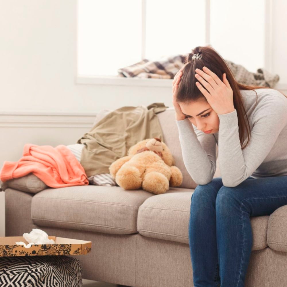 A young woman sits on a couch with her head in her hands, looking distressed. The sofa behind her is messy with clothes and a teddy bear, and a pizza box with tissues sits on the table in front of her.