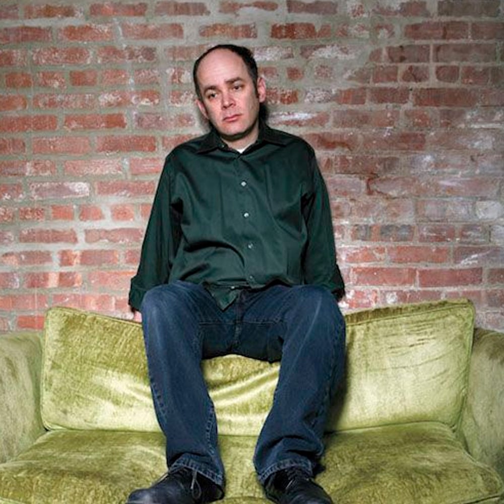 A man in a dark green shirt and jeans sits on a light green velvet couch against an exposed brick wall, looking slightly pensive with his hands resting on the seat.