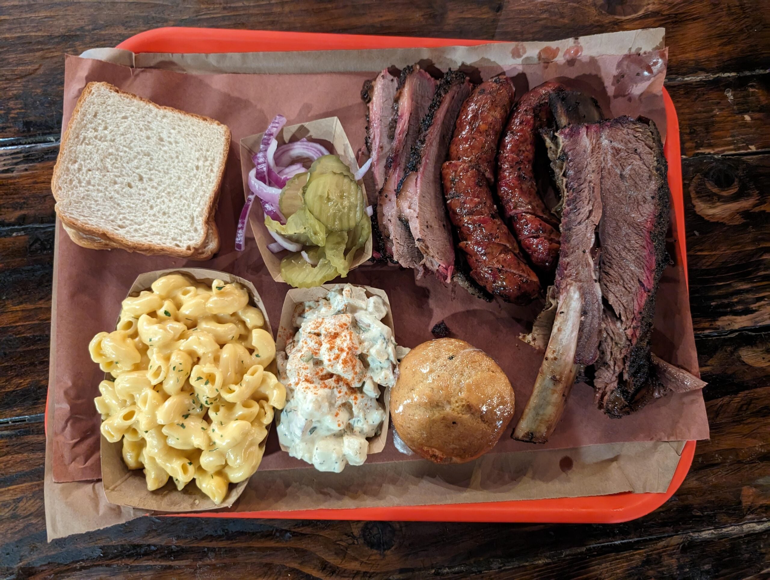A tray of barbecue food with sliced brisket, sausage, macaroni and cheese, potato salad, a bread slice, a muffin, pickles, and onion slices on brown paper.