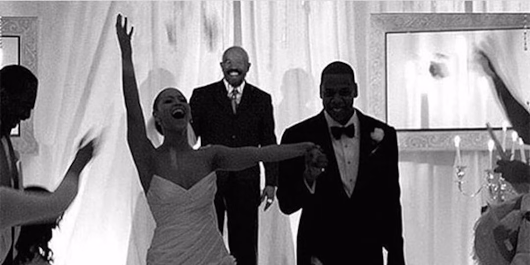 A bride and groom celebrate, holding hands and smiling, as guests cheer around them at their wedding. The couple is dressed formally, with the bride in a gown and the groom in a tuxedo. Curtains and candles decorate the background.
