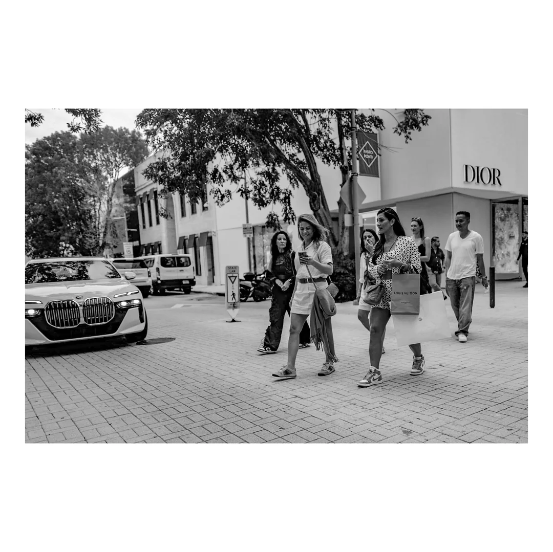 A group of people walks across a city street near a Dior store, with a car approaching from the left and trees and buildings in the background. The image is in black and white.