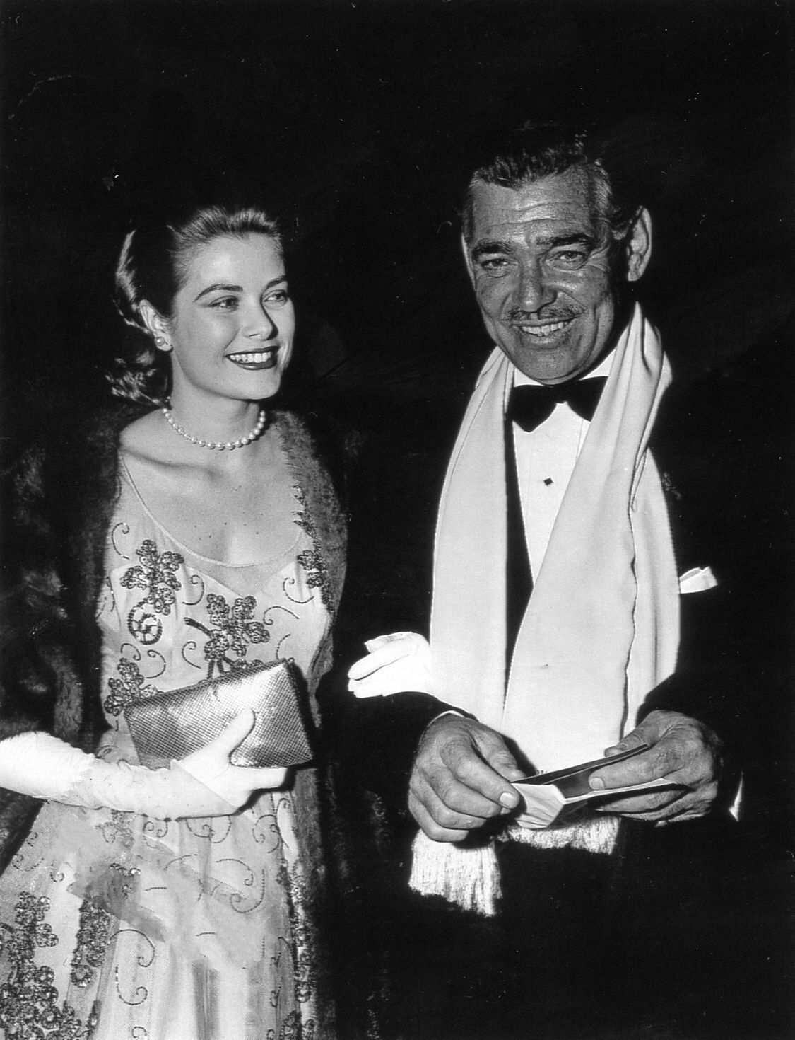 A woman in an embellished gown and gloves smiles beside a man in a tuxedo and scarf. Both appear dressed for a formal event, standing close together and looking happy. The photo is in black and white.