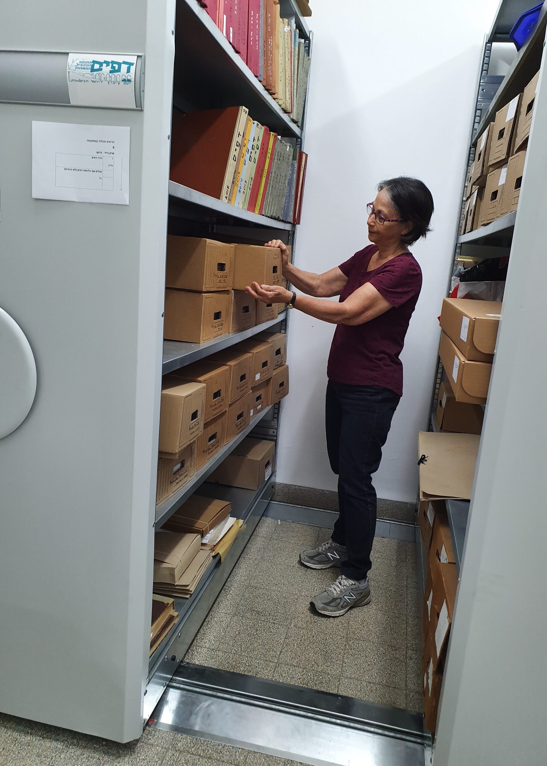 A woman wearing glasses, a maroon shirt, and dark pants is standing in a storage room, organizing brown archive boxes on metal shelves filled with files and documents.