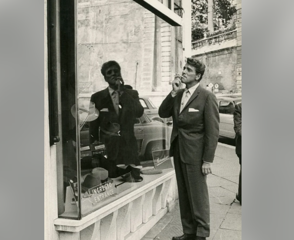 A man in a suit stands on a sidewalk, looking thoughtfully into a shop window with his hand on his chin. His reflection is visible in the glass, and vintage cars are parked on the street behind him.