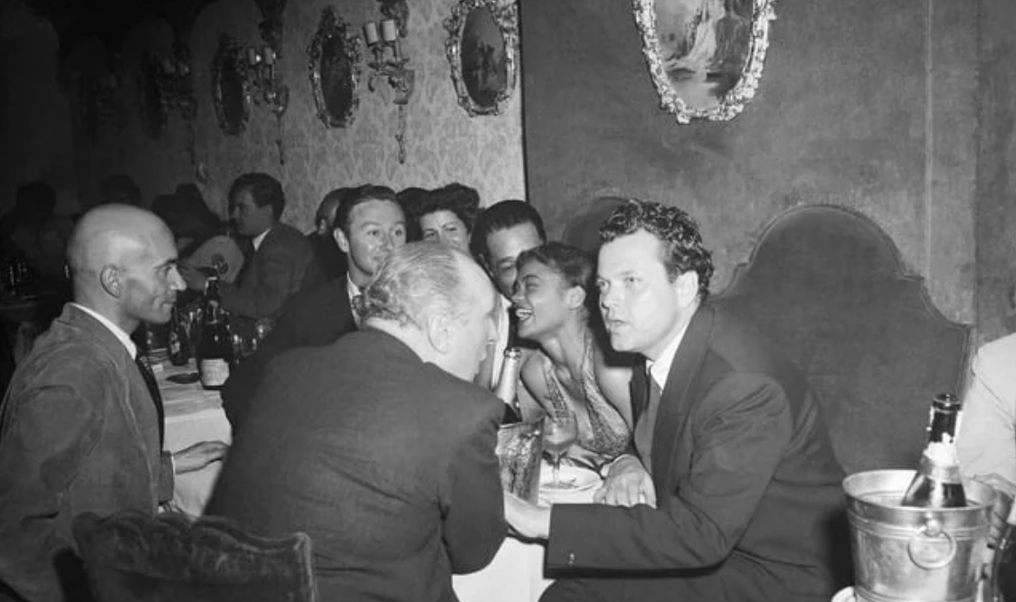 A group of people in formal attire sit around a crowded table at a dimly lit restaurant or club, engaged in conversation, with bottles and glasses on the table and ornate mirrors on the wall behind them.