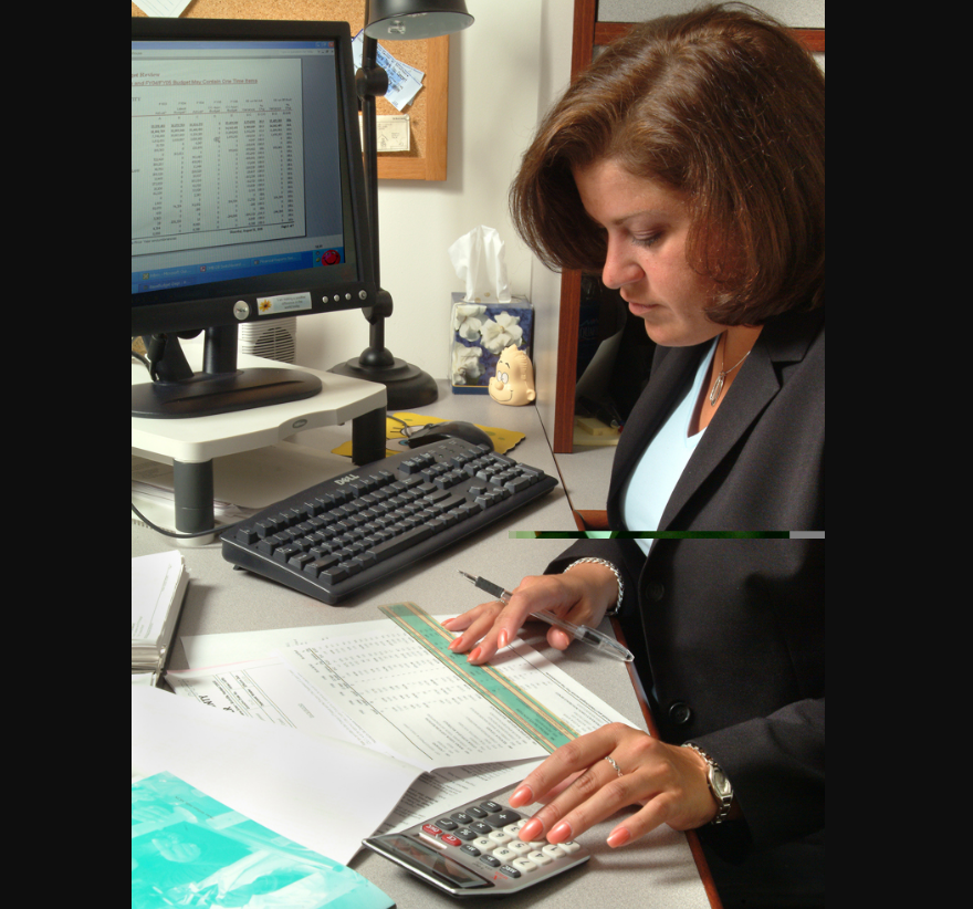A woman in business attire sits at a desk, using a calculator and reviewing documents. A computer monitor with a spreadsheet is on the desk, along with a keyboard, mouse, and office supplies.