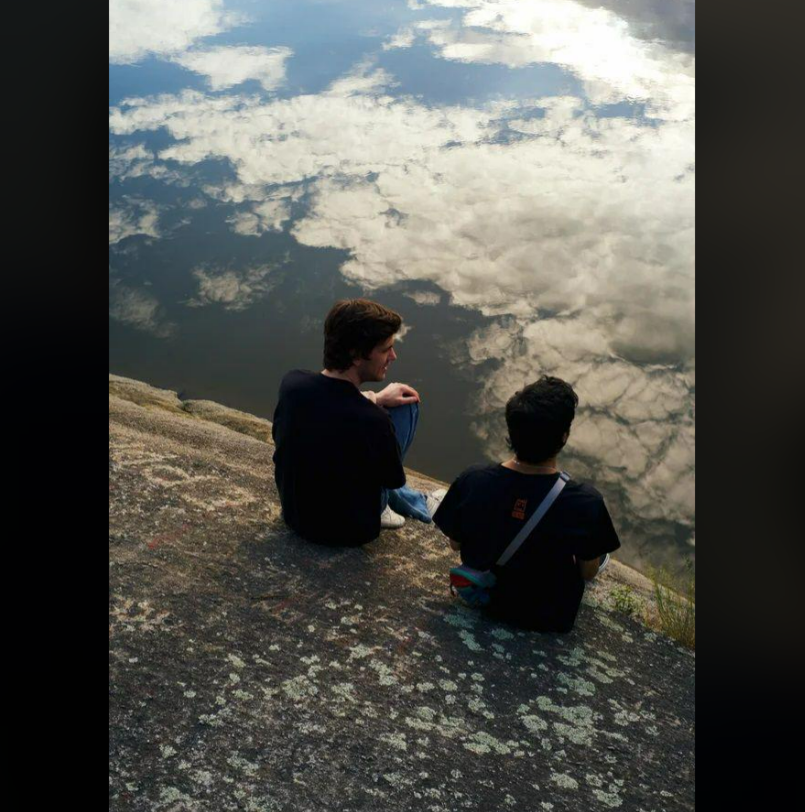Two people sit on a rocky surface beside a body of water, which beautifully reflects the sky and clouds above them, creating a serene and contemplative scene.