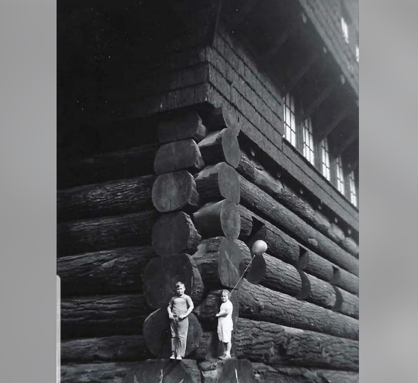 Two children stand on large logs at the corner of a rustic log cabin, with one child holding a balloon. The building is tall with layered log construction and several windows above. The image is in black and white.