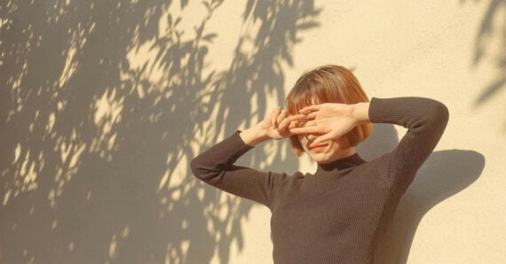 A person with short hair wearing a black long-sleeve top stands against a sunlit wall, shielding their eyes from the bright light with both hands. Leaf shadows are cast on the wall around them.