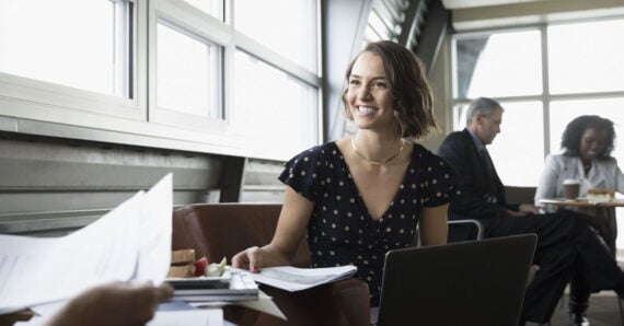 A woman in a polka-dot dress smiles while sitting at a table with papers and a laptop. Two people converse in the background in a bright, modern office or caf&eacute; setting.